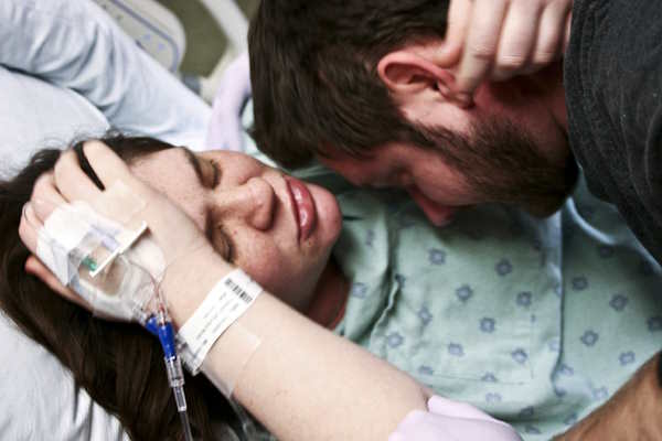 Fotografia a colori di una paziente sdraiata su un letto d'ospedale che indossa un camice da ospedale e si tiene la testa tra le mani.